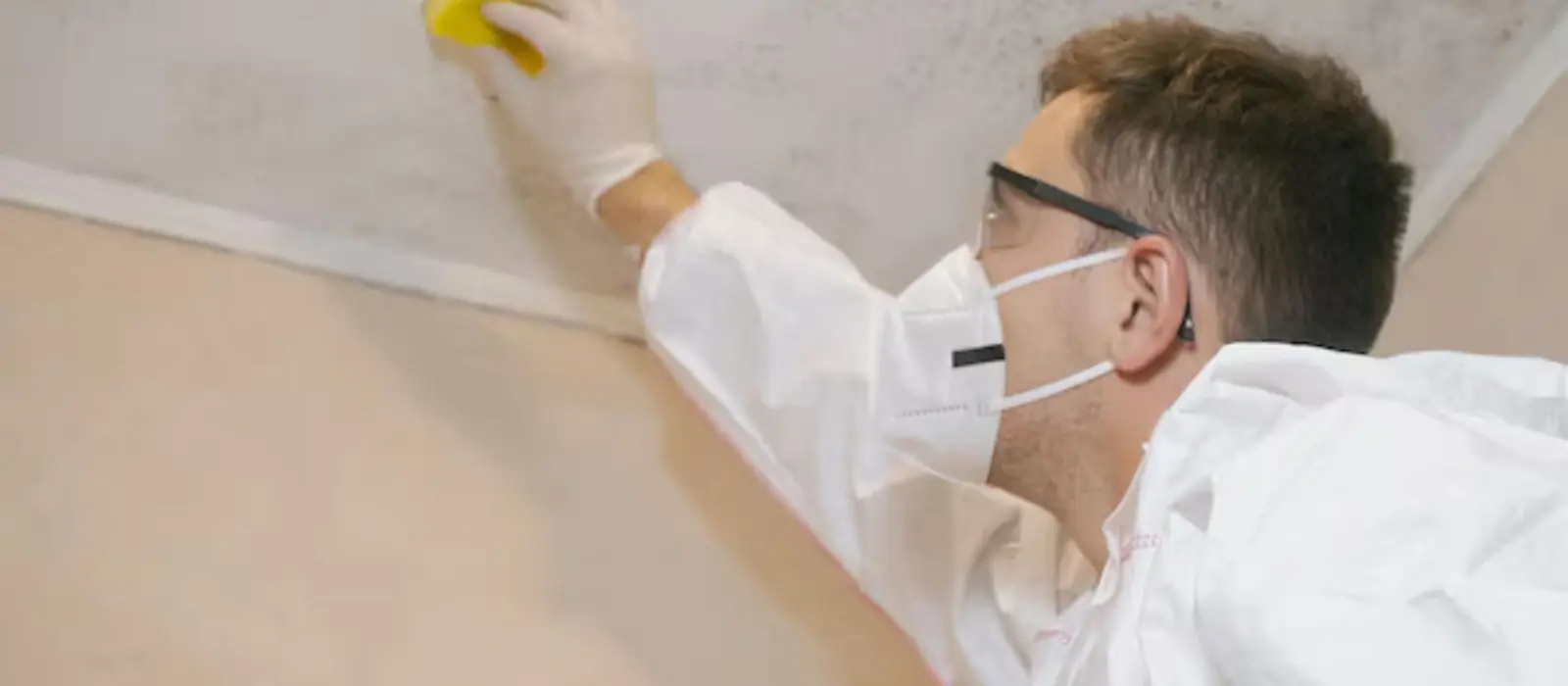Person in a mask and protective suit wiping mold from a ceiling corner
